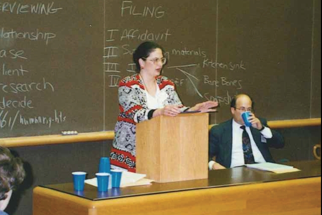 [ai] A woman speaking at a podium in front of a chalkboard with notes. A man sitting next to her is holding a cup. Blue cups are placed on the table in front. The setting appears to be a classroom or seminar room.